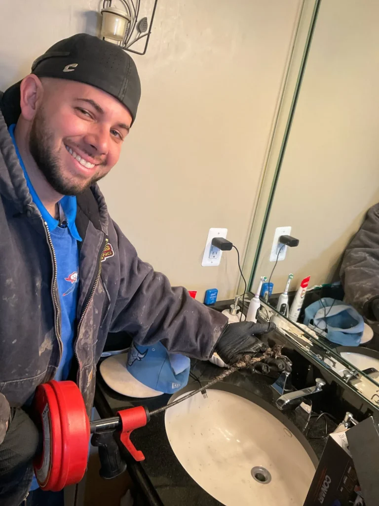 A smiling person holds plumbing tools and debris over a bathroom sink, ready for some serious drain clearing. Wearing a cap and jacket, their reflection in the mirror reveals wall outlets and personal items scattered on the counter.