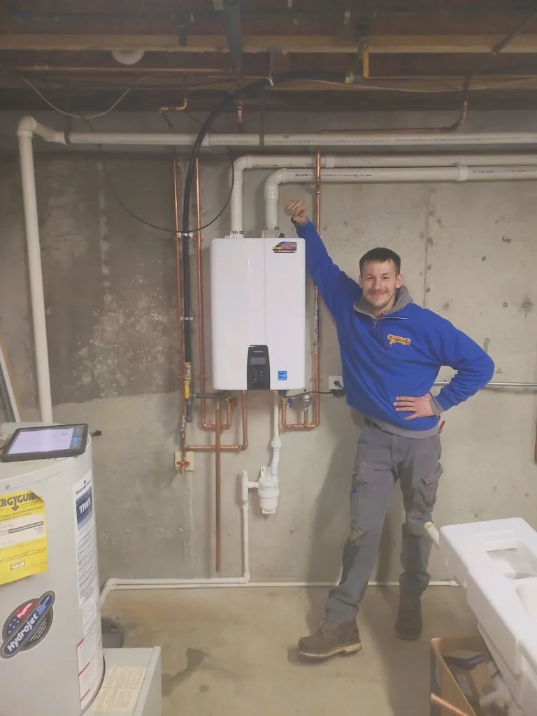 A person in a blue sweatshirt stands next to a wall-mounted tankless water heater in a basement, gesturing proudly. Pipes and HVAC-related equipment are visible around them.