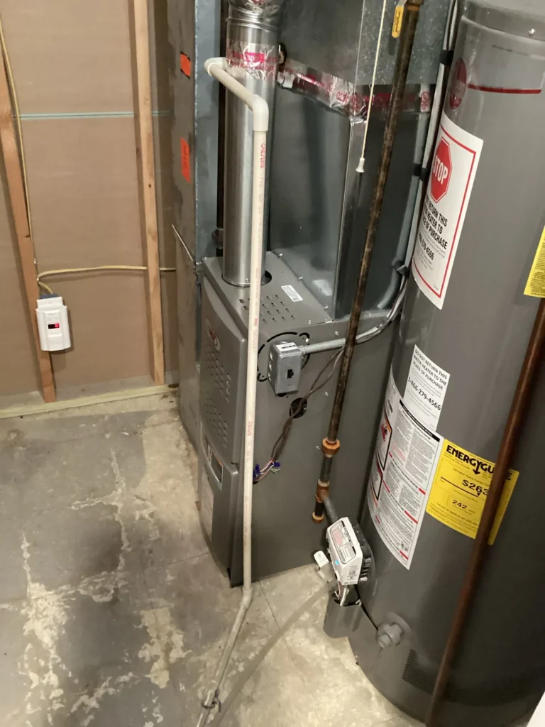 Basement utility area with a furnace and water heater. PVC pipes connect to the units, and an electrical outlet is nearby. Walls are unfinished, showing wooden studs.