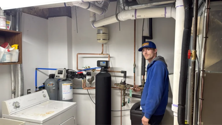 A person in a blue hoodie and cap adjusts equipment in a utility room with pipes, a water softener, and a washer.