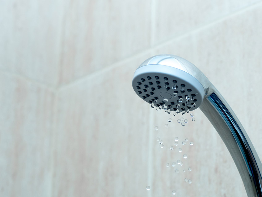 Close-up of a showerhead with water droplets emerging, against a light stone-tiled background.